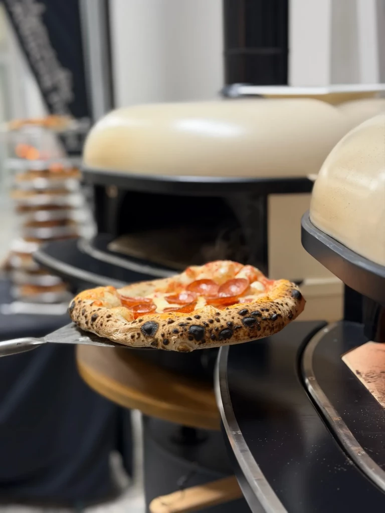 Frisch gebackene Pizza Salame auf dem Pizzaschieber vor zwei mobilen Pizzaöfen