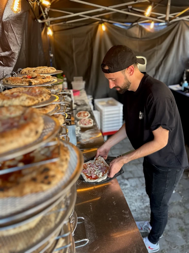 Pizzabäcker belegt eine Pizza am Stand, daneben fertige Pizzen auf Etageren